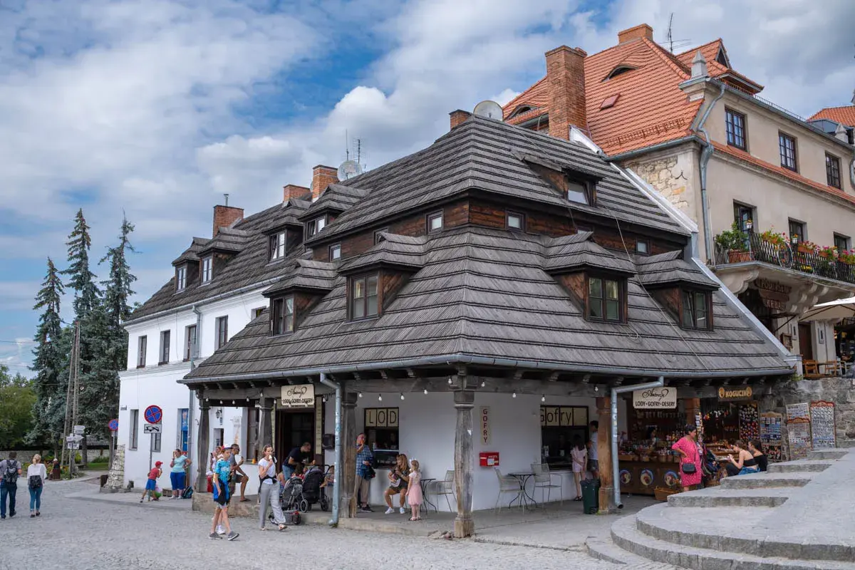 Tętniący życiem rynek w Kazimierzu Dolnym. Ludzie spacerują, kupują lody i gofry pod charakterystycznymi drewnianymi dachami.