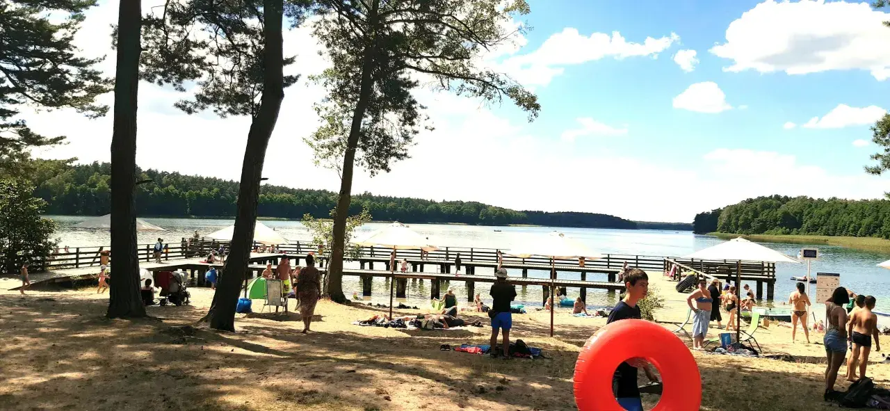 Słoneczny dzień nad jeziorem. Ludzie odpoczywają na plaży i molo, ciesząc się wakacjami. W tle las i błękitne niebo.