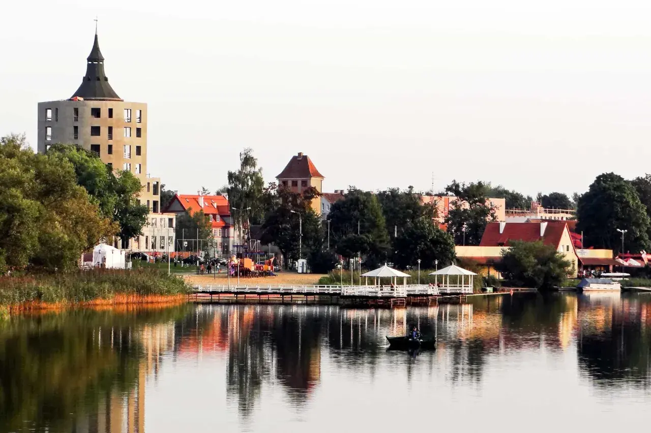 Widok na jezioro z łodzią, pomostem z altanami i budynkami w tle.