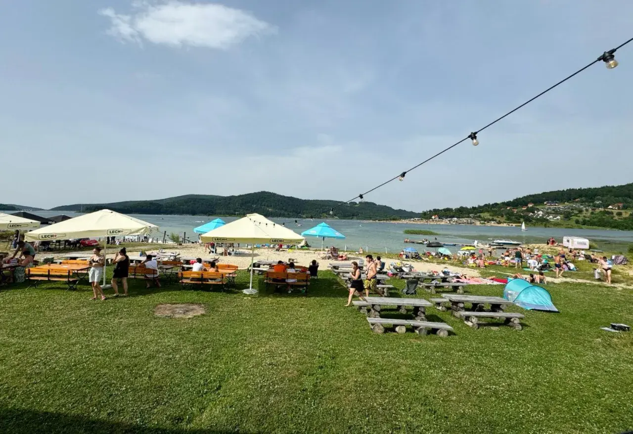 Plaża nad jeziorem z ludźmi, parasolami, stolikami i ławkami w słoneczny dzień.