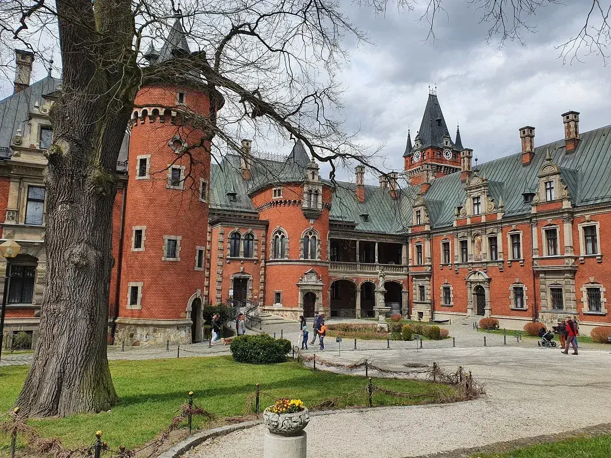Pławniowice atrakcje: ceglany zamek z wieżą i zegarem, dziedziniec z drzewem i ludźmi spacerującymi po okolicy.