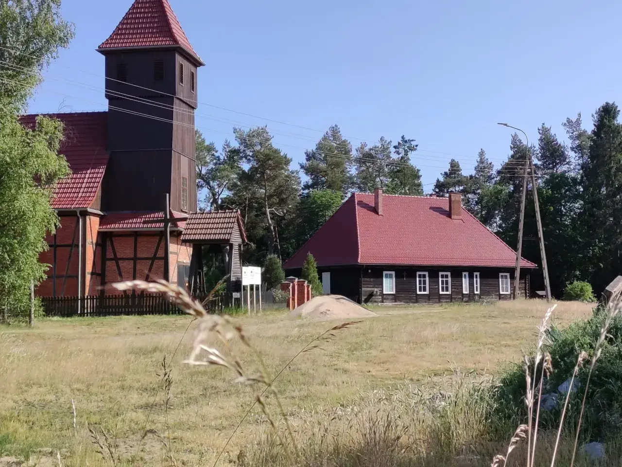 Drewniana wieża kościelna i dom z czerwonym dachem w Starowej Górze, otoczone polami i drzewami.