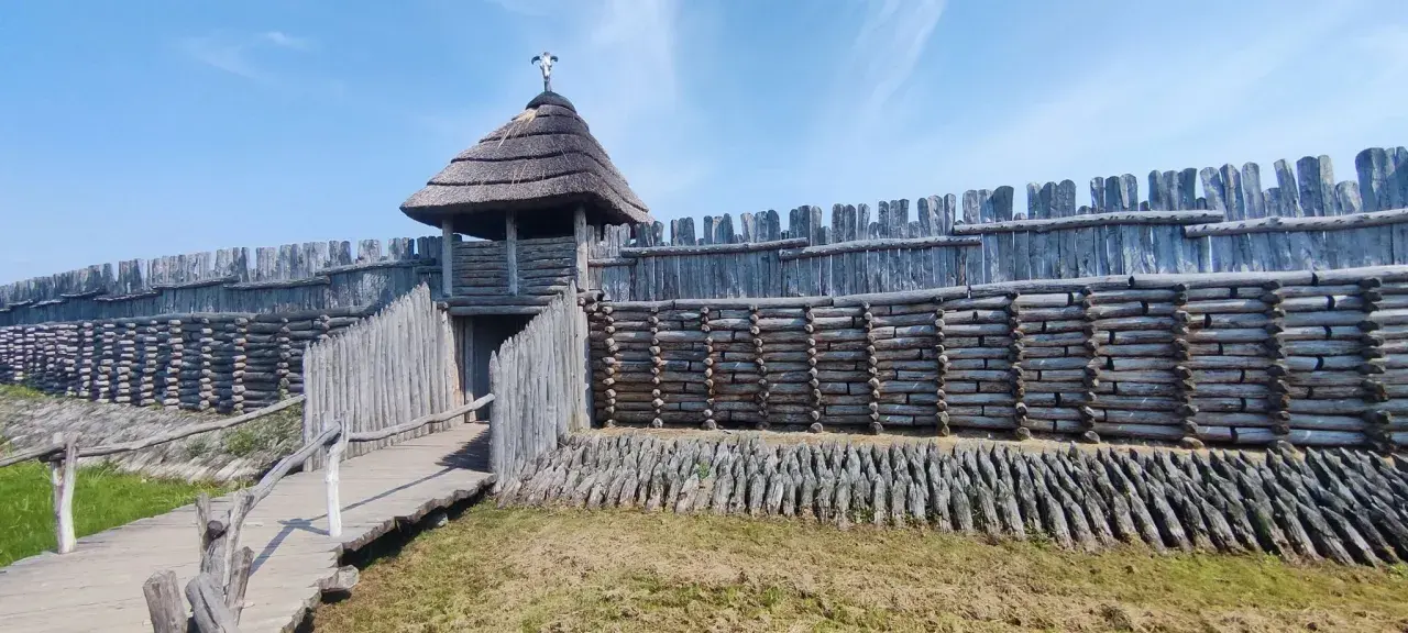 Drewniany gród obronny z wieżą i strzechą, pod niebieskim niebem.