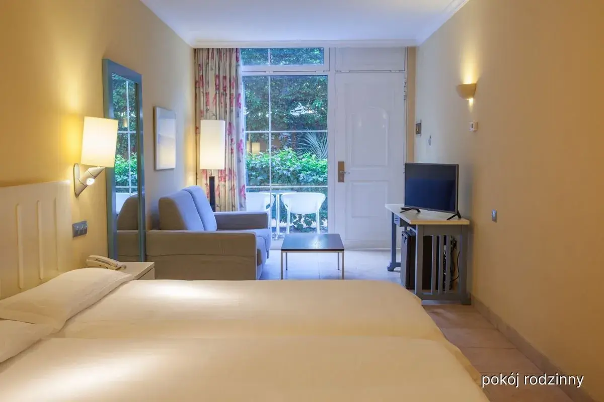 Pokój rodzinny w hotelu IFA Altamarena: łóżko, sofa, TV i wyjście na taras z widokiem na zieleń.