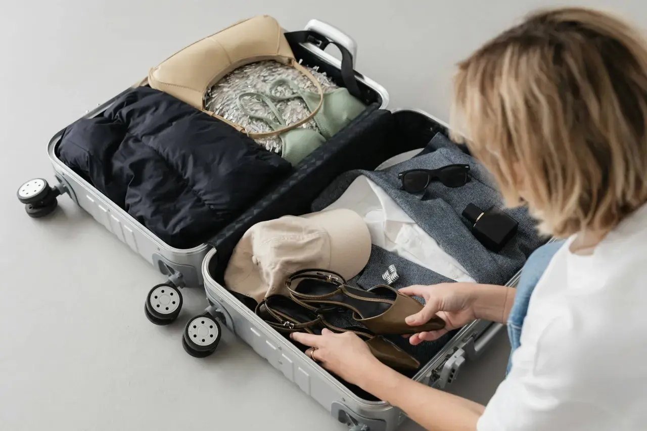 Kobieta pakuje do walizki: czarną kurtkę, zielony komplet, beżową torebkę, czapkę, buty, okulary i perfumy. Gotowa na podróż z enter air bagaż.