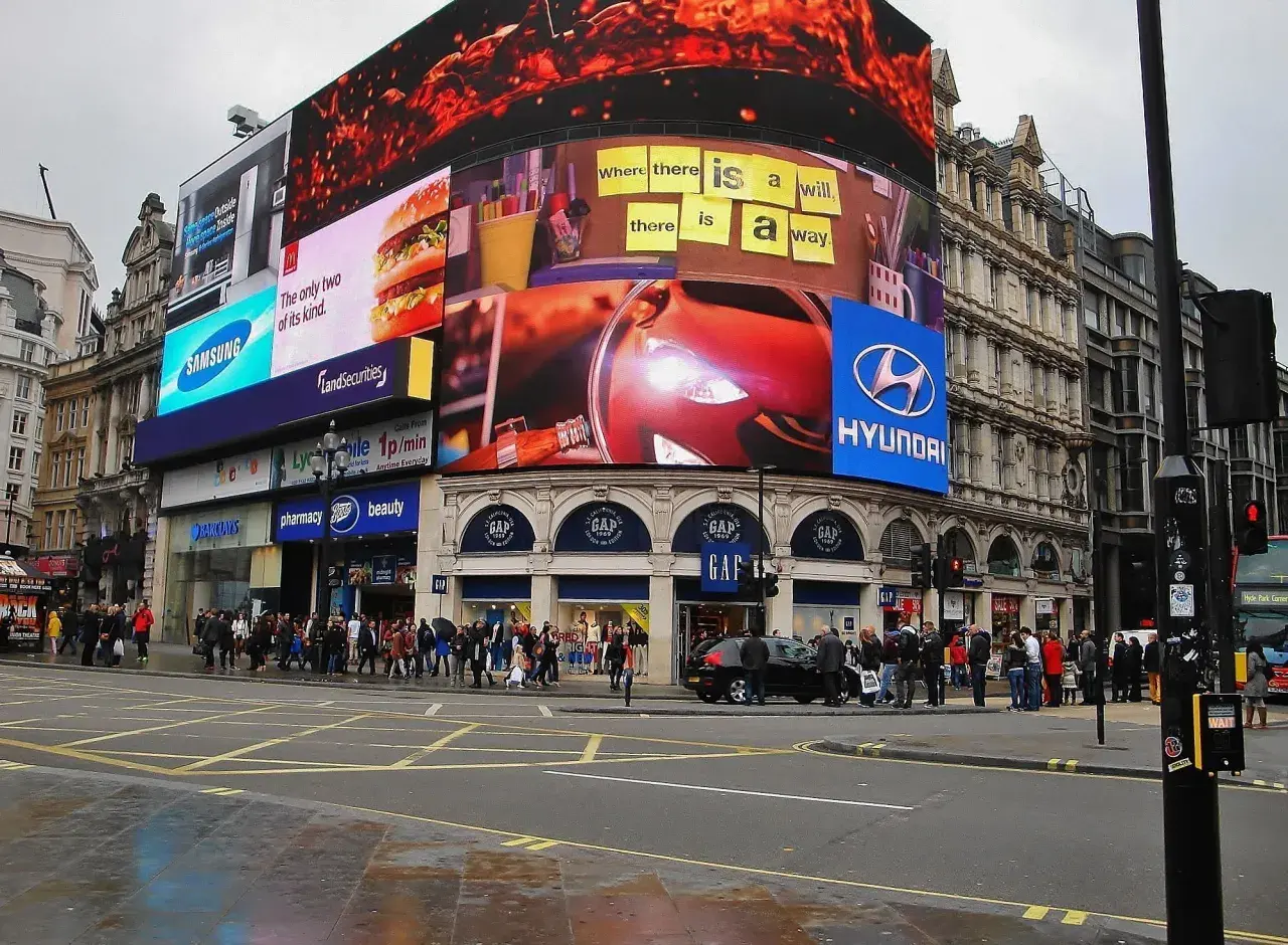 Tętniący życiem Piccadilly Circus w Londynie z neonowymi reklamami, w tym Samsung, McDonald's i Hyundai. Tłum ludzi na chodniku.