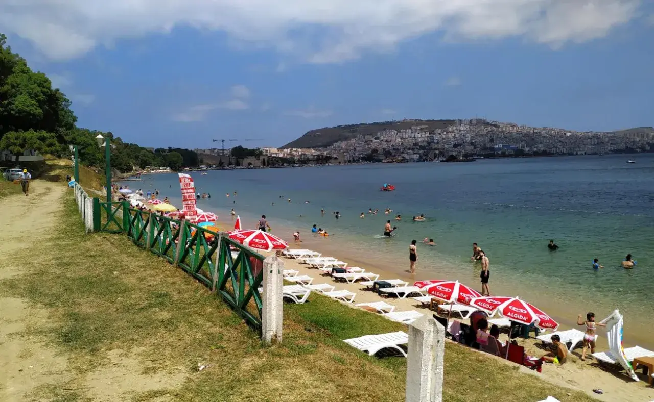 Plaża z leżakami, parasolami i ludźmi kąpiącymi się w morzu, w tle miasto na wzgórzu.