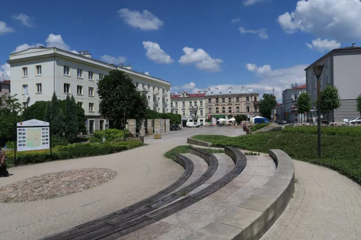 Słoneczny dzień na placu artystów w Kielcach. Widać budynki, zieleń i ławki.