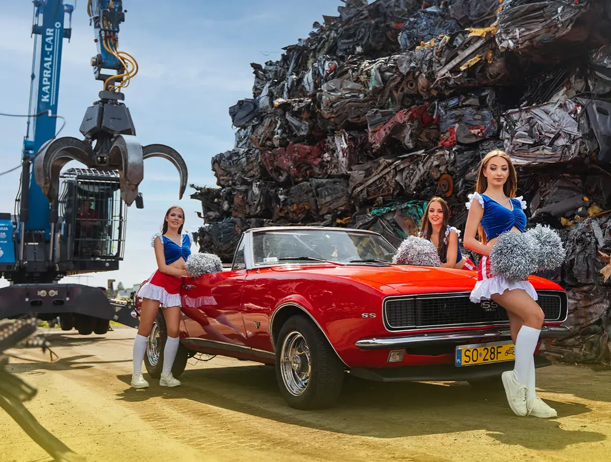 Trzy cheerleaderki w czerwono-niebieskich strojach stoją obok czerwonego kabrioletu na tle złomowiska.