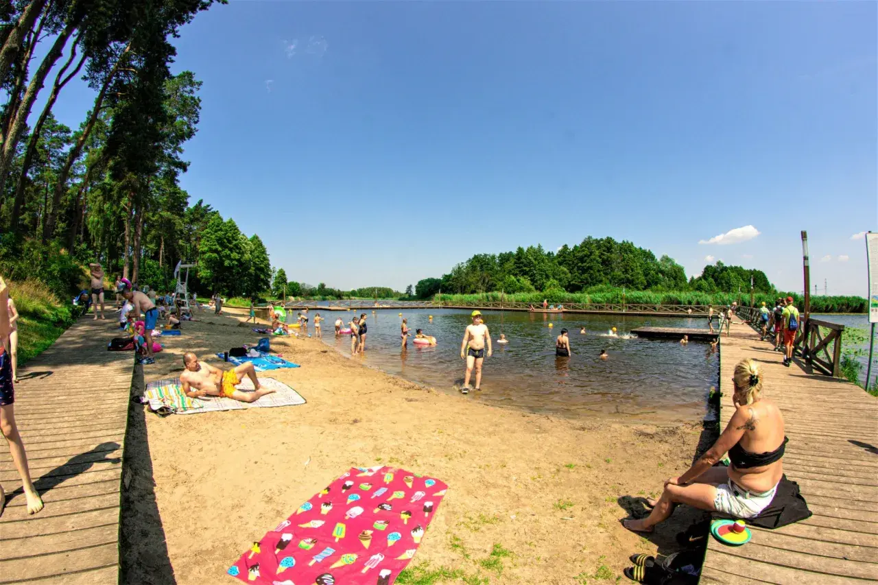Ludzie odpoczywają i kąpią się nad **jeziorem Kozienickim** w słoneczny dzień.