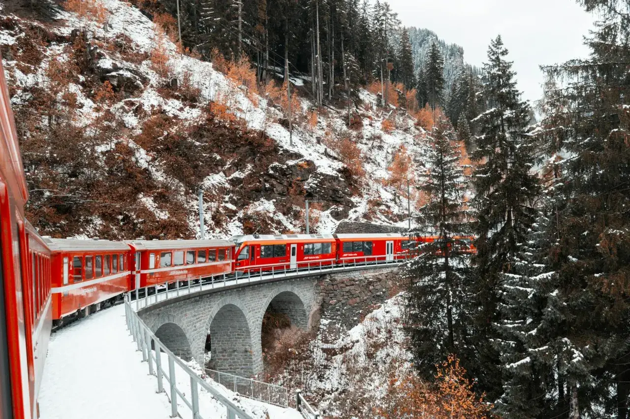 Czerwony pociąg Glacier Express przejeżdża przez kamienny most w zimowym, zaśnieżonym krajobrazie górskim.