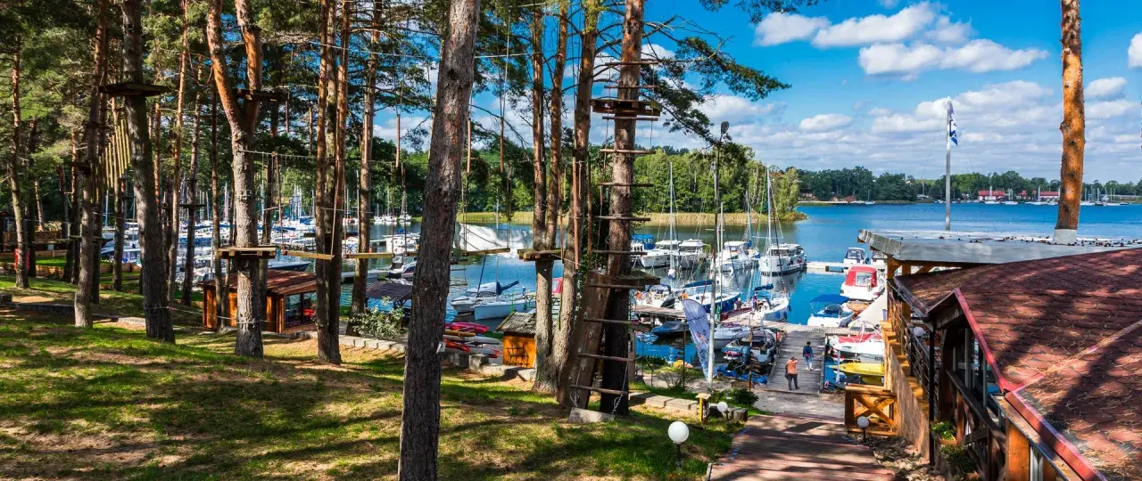 Linowy park rozrywki w lesie nad jeziorem z przystanią pełną jachtów.