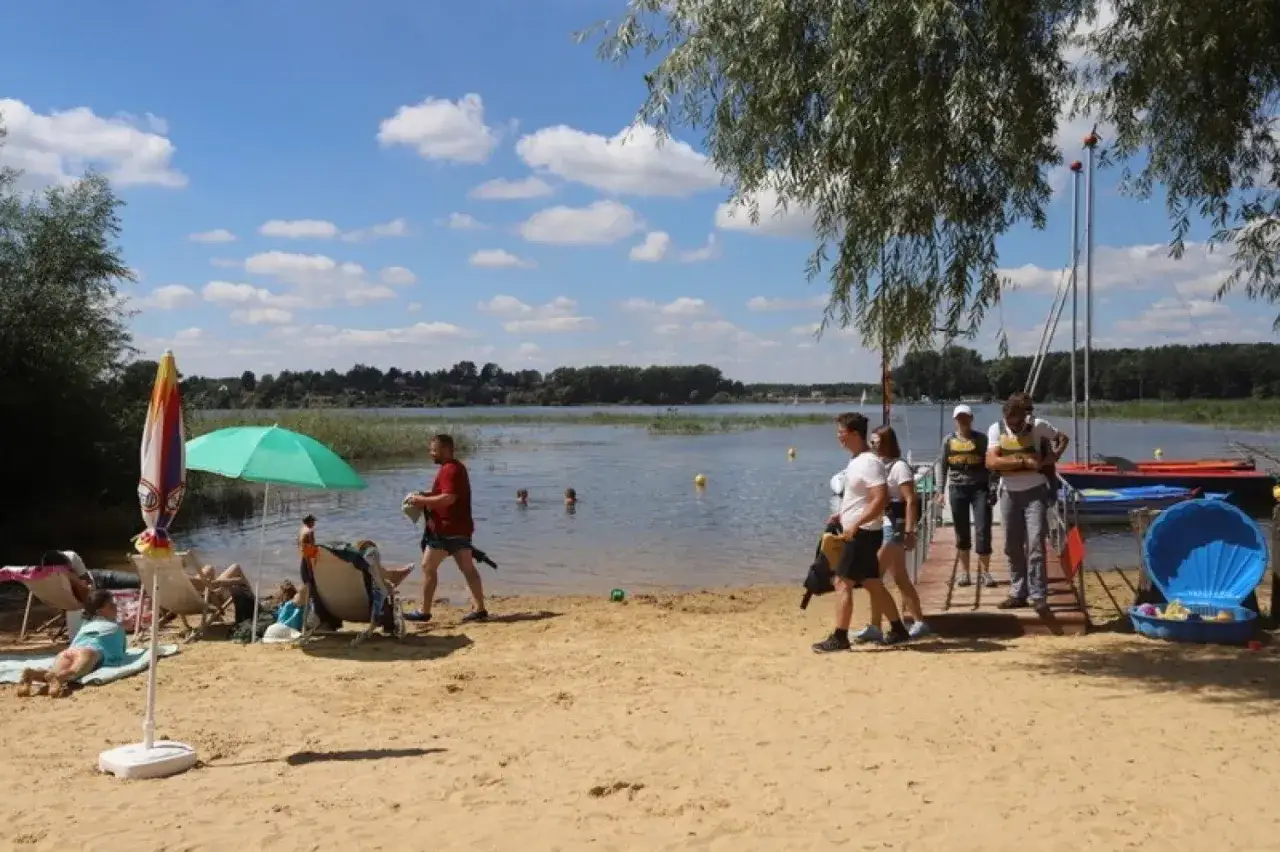 Na plaży nad jeziorem przeczyckim widać ludzi odpoczywających i kąpiących się. W tle żaglówki i drzewa.