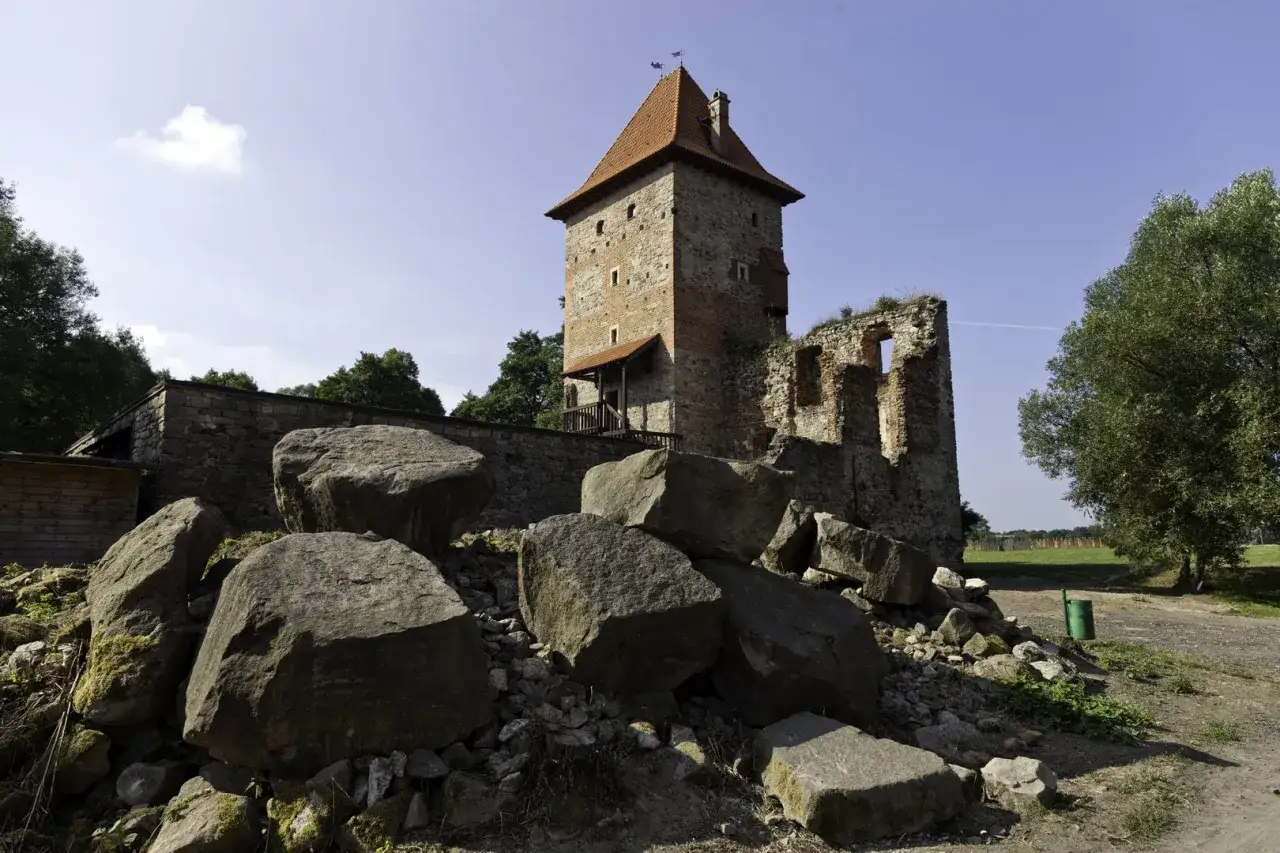 Kamienny zamek w Chudowie, ruiny i wieża z czerwonym dachem. Wokół wielkie głazy, zielone drzewa i błękitne niebo. Chudów atrakcje dla miłośników historii.