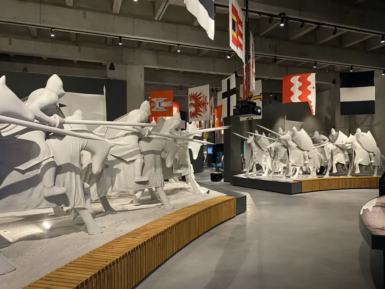 Białe, stylizowane figury rycerzy na koniach, przypominające scenę bitwy. W tle wiszą historyczne flagi. To jedne z wielu atrakcji w muzeum Grunwald.