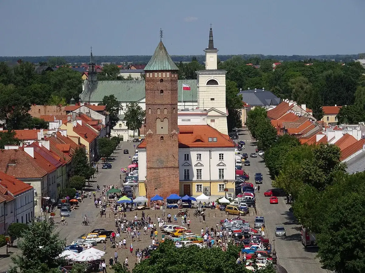 Tętniący życiem rynek w Pułtusku z wieżą zamkową, kościołem i kolorowymi kamieniczkami. Ludzie spacerują, oglądają samochody i stoiska.