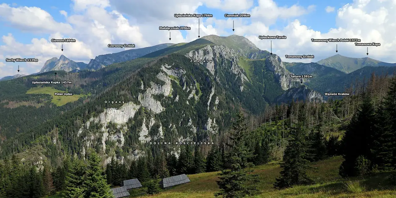Widok na Tatry z Doliną Kościeliską. W oddali majaczą szczyty, w tym Giewont. Na pierwszym planie widać drewniane hale, stoły pasterskie.