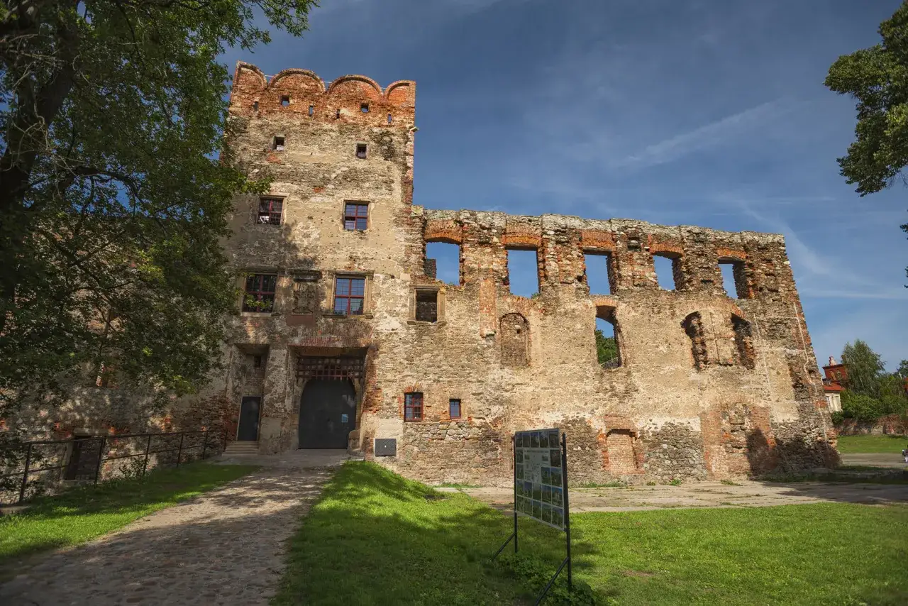 Zamek w Ząbkowicach Śląskich, ruiny z ceglanymi detalami i wieżą, otoczone zielenią i brukowanym podjazdem.