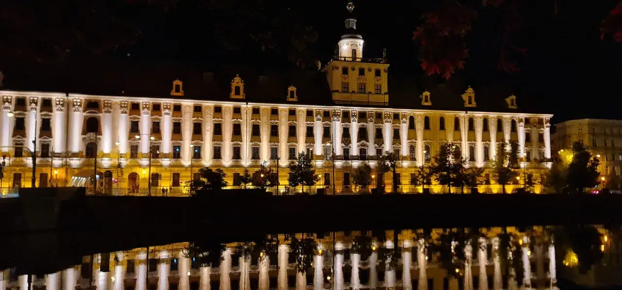 Piękny, nocny widok na Uniwersytet Wrocławski, który znajduje się na placu solnym we Wrocławiu. Budynek jest oświetlony, a jego odbicie w wodzie tworzy magiczny obraz.
