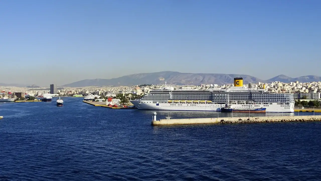 Ogromny statek wycieczkowy cumuje w porcie Piraeus, z widokiem na miasto i góry w tle.