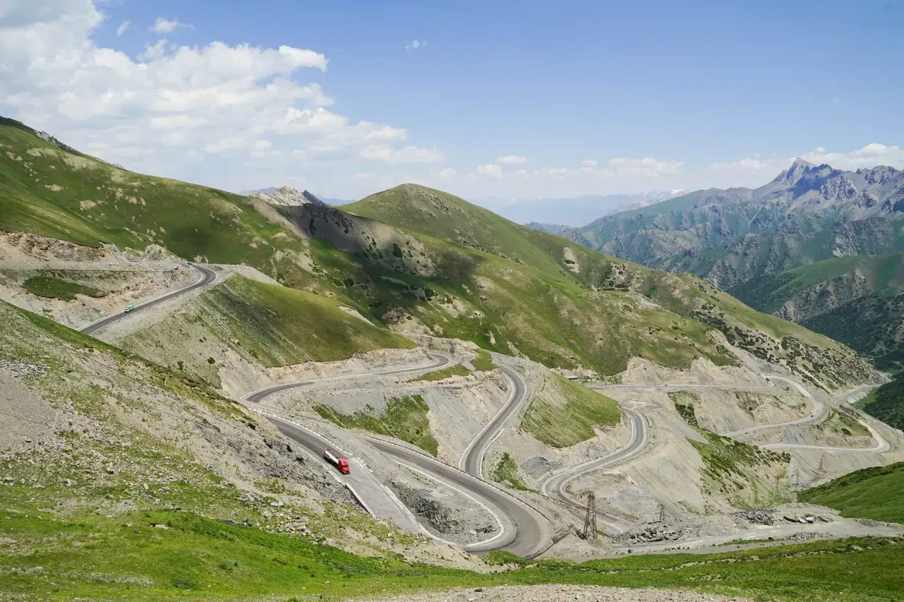 Kręta droga na Pamir Highway wijąca się po zielonych zboczach gór. Widać ciężarówkę i samochód.