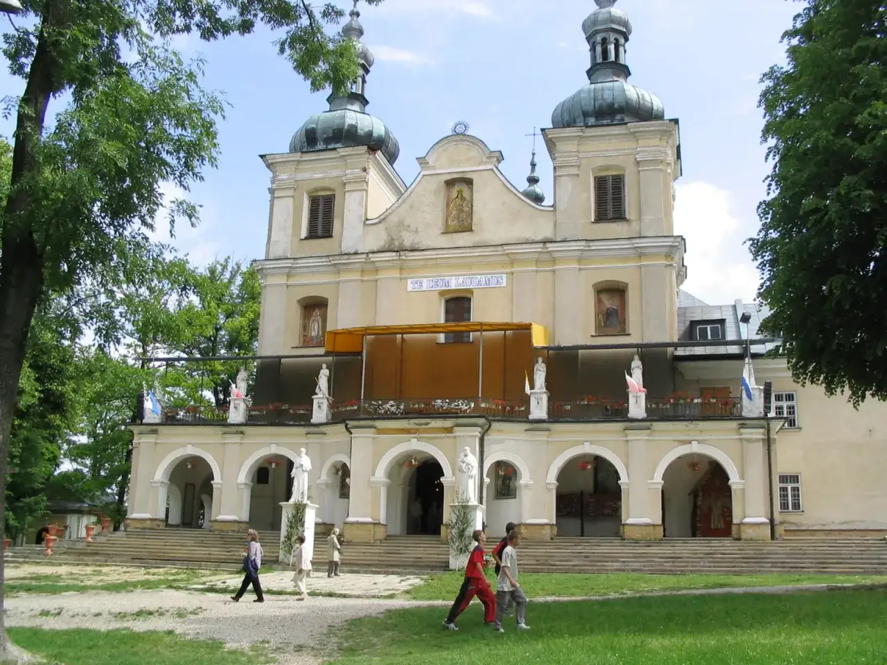 Barokowa bazylika z kopułami, otoczona zielenią. Wokół kościoła spacerują ludzie, odkrywając Arłamów atrakcje i piękno tego miejsca.