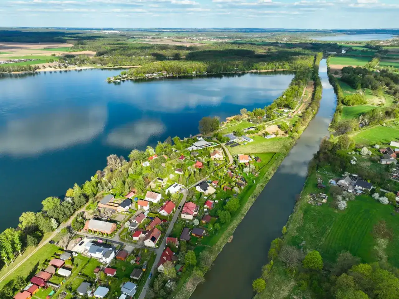 Malownicze jezioro Pławniowice otoczone zielenią i domkami letniskowymi, z kanałem wodnym biegnącym wzdłuż brzegu.