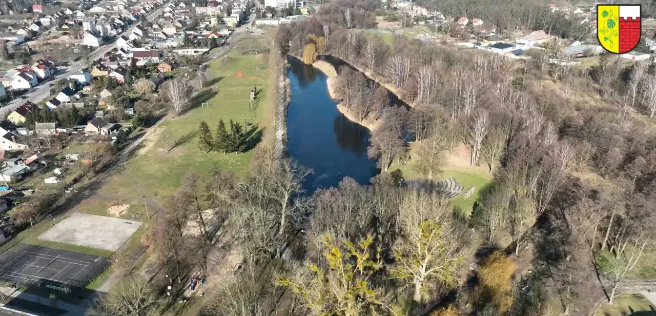 Widok z lotu ptaka na miasto i jezioro. W oddali widać zabudowania, a bliżej tereny zielone z boiskiem i kortami tenisowymi. W lesie ukrywa się je jeleń witnica.