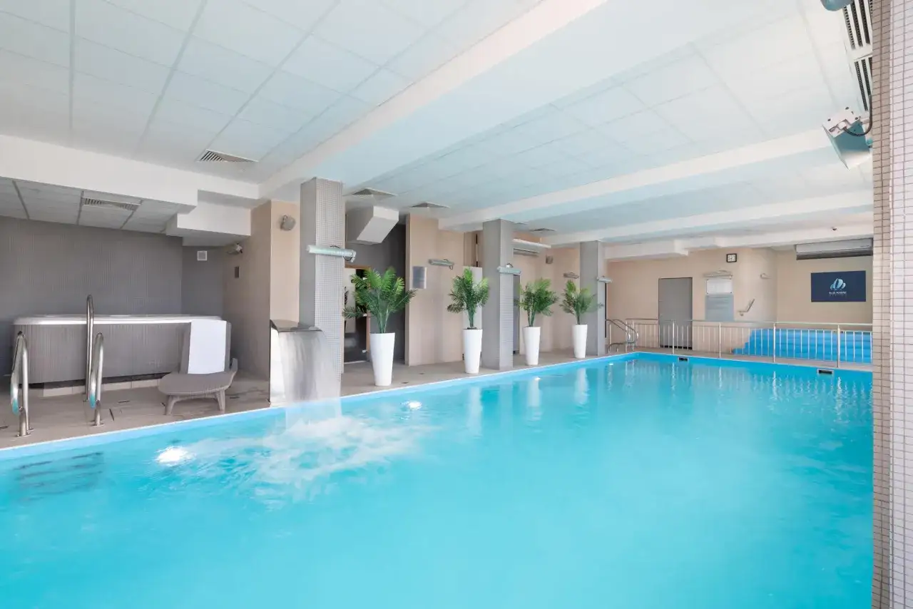 Basen z wodospadem i jacuzzi w hotelu Blue Marine. Wnętrze z palmami w białych donicach i leżakiem.