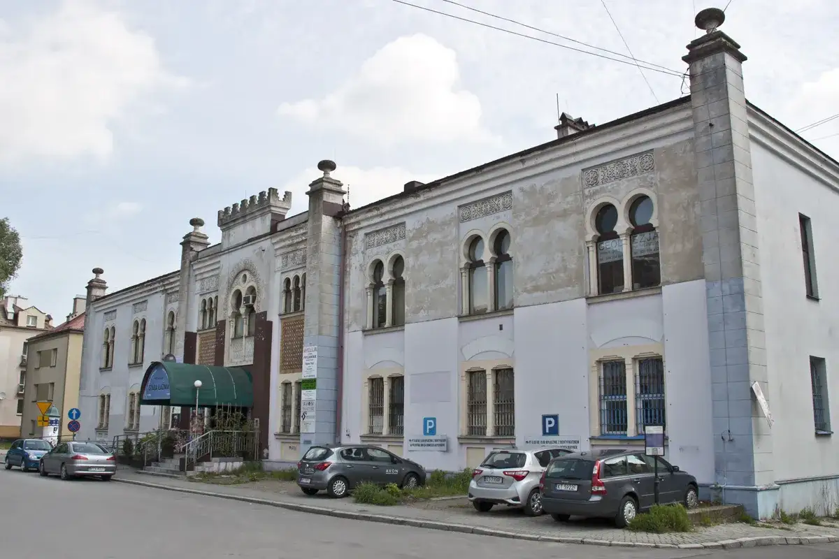 Elegancka fasada Starej Łaźni w Tarnowie z charakterystycznymi arkadowymi oknami. Przed budynkiem zaparkowane samochody.
