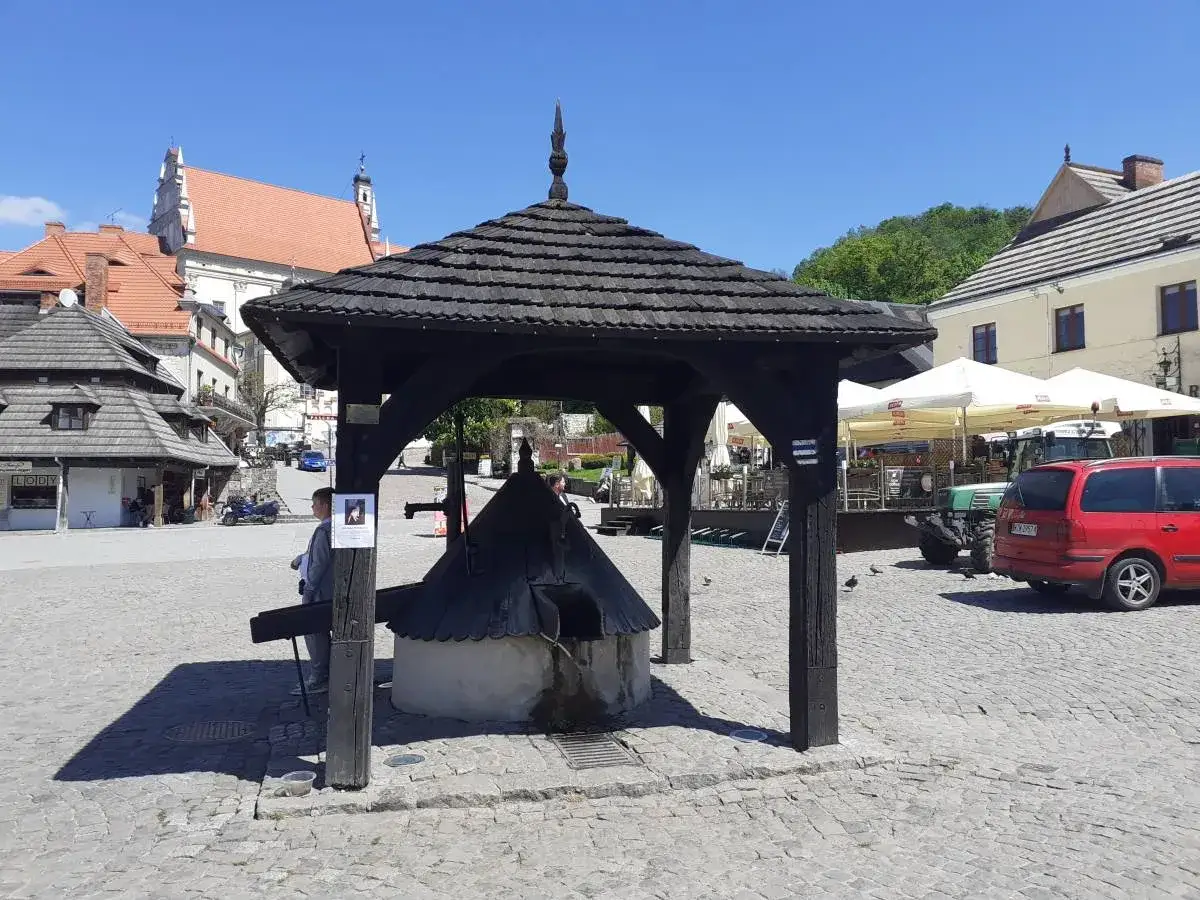 Drewniana studnia na brukowanym rynku w Kazimierzu Dolnym. W tle kamienice i kościół.