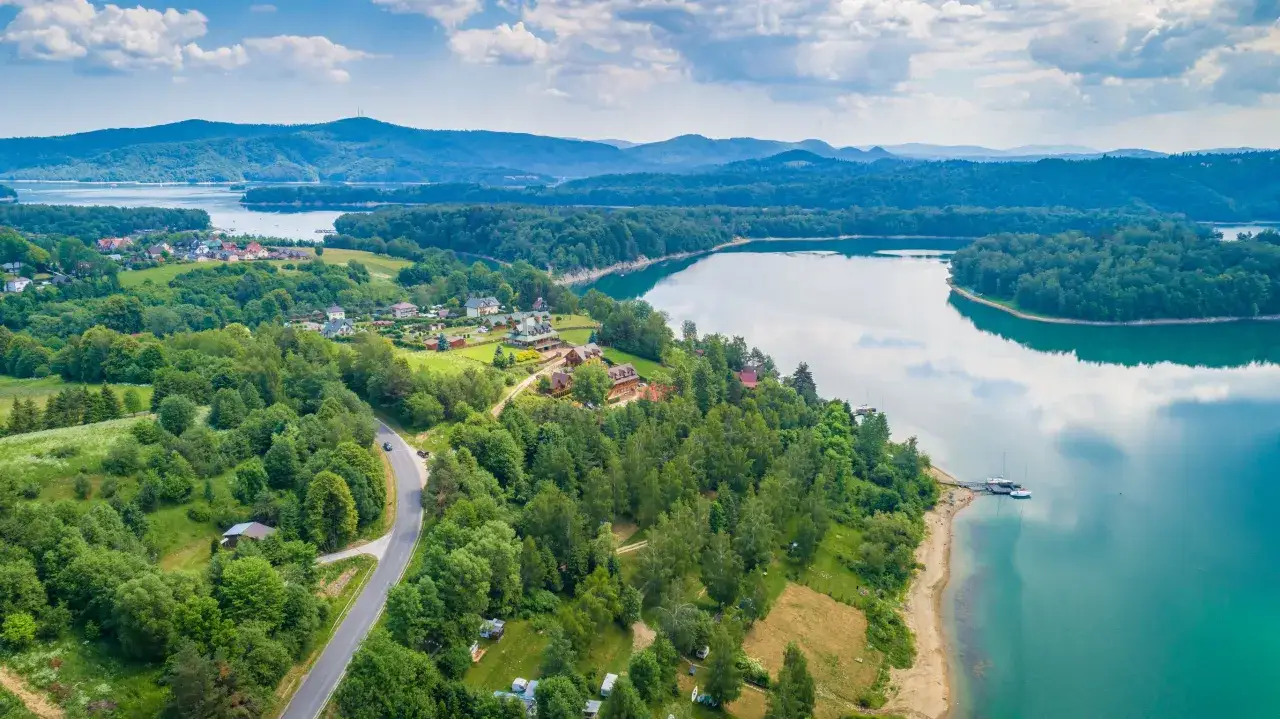 Malownicze widoki na Jezioro Solińskie i okoliczne wzgórza. Widać domki letniskowe, lasy i drogę. Idealne miejsce na odkrywanie atrakcji Solina i okolice.